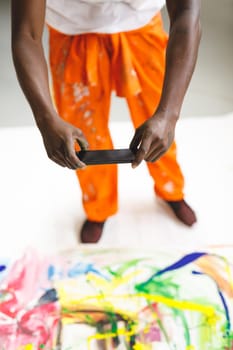 African american male painter at work taking picture of artwork with smartphone in art studio. creation and inspiration at an artists painting studio.