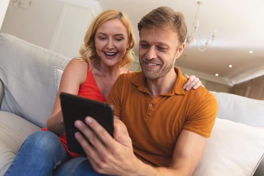 Caucasian couple sitting on couch smiling and using tablet at home. enjoying free time relaxing with technology at home.