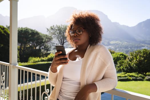 Happy african american woman standing on sunny balcony of country home using smartphone. domestic lifestyle, enjoying leisure time at home.