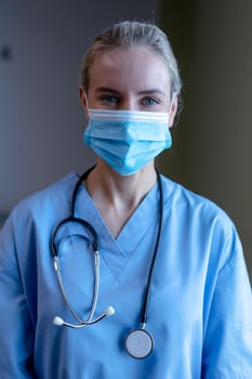 Portrait of caucasian female doctor wearing face mask, scrubs and stethoscope looking to camera. medical professional at work and health services during coronavirus covid 19 pandemic.