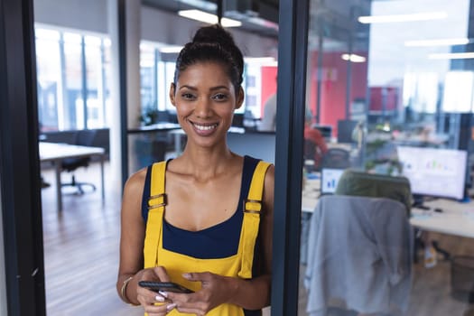 Portrait of mixed race female creative worker using smartphone and smiling. modern office of a creative design business.