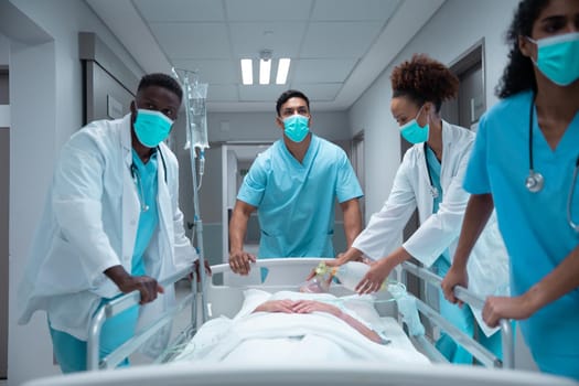 Mixed race doctors wearing face masks transporting patient in hospital bed. medicine, health and healthcare services during coronavirus covid 19 pandemic.