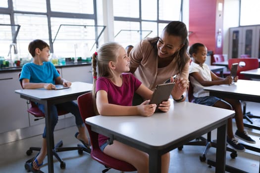 African american female teacher teaching caucasian girl to use digital tablet in the class at school. school and education concept