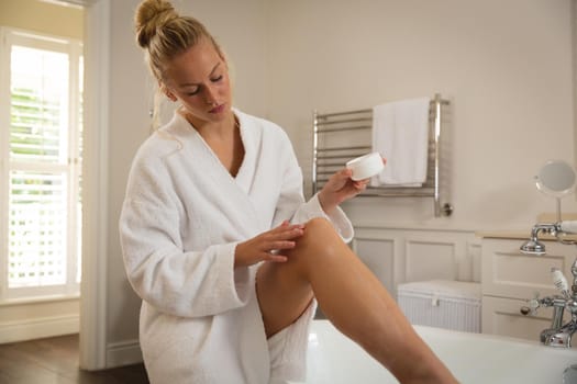 Caucasian woman sitting in bathroom wearing bathrobe moisturising legs. health, beauty and wellbeing, spending quality time at home.