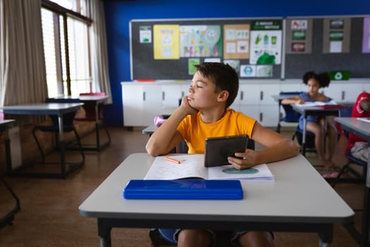 Caucasian boy holding digital tablet while sitting on the desk in the class at elementary school. school and education concept