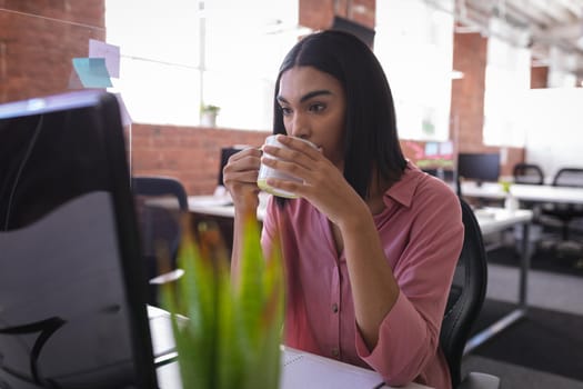 Mixed race businesswoman sitting in office in front of computer and having coffee. independent creative design business.