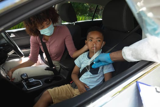 Medical worker wearing ppe suit taking swab tests of african american mother with son sitting in car. medical precautions, lifestyle during covid 19 pandemic.