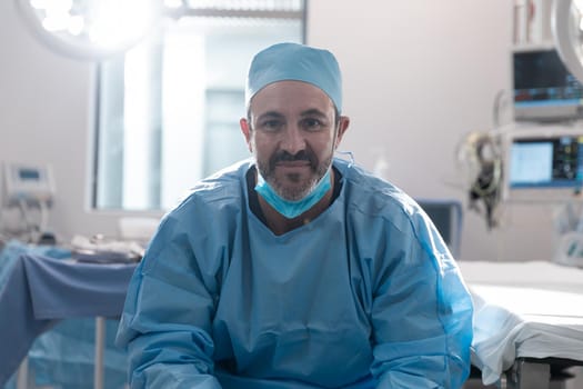 Smiling caucasian male surgeon with face mask wearing protective clothing in operating theatre. medicine, health and healthcare services during covid 19 coronavirus pandemic.