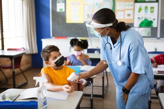 Female health worker wearing face shield measuring temperature of a boy at elementary school. education back to school health safety during covid19 coronavirus pandemic.