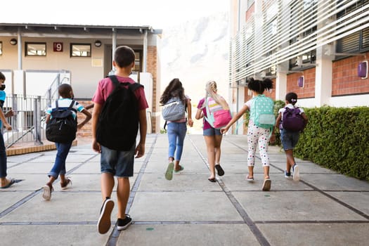 Rear view of group of diverse students with backpacks running at elementary school. education back to school health safety during covid19 coronavirus pandemic.