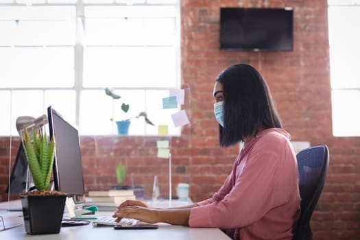 Mixed race businesswoman sitting in office in front of computer wearing face mask. independent creative design business during covid 19 coronavirus pandemic.