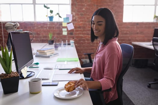 Mixed race businesswoman sitting in office in front of computer and having snack. independent creative design business.