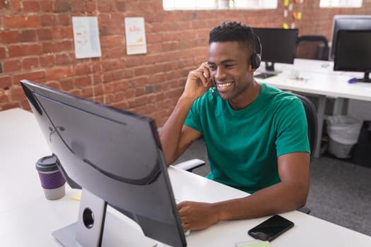 African american businessman having video call sitting in front of computer using headphones. independent creative design business.