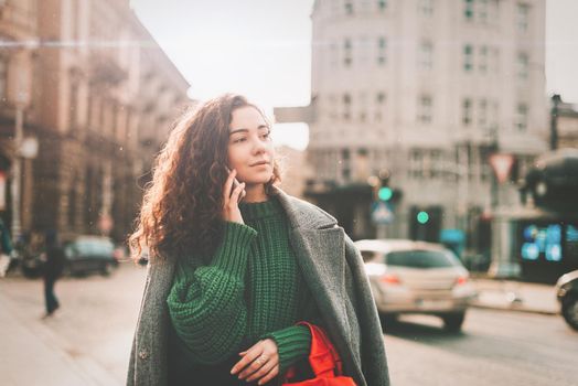 A woman on the street uses a mobile phone. online shopping. use of mobile applications. beautiful young woman with long curly dark hair in a casual coat, trendy green sweater and red handbag