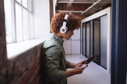 Portrait of happy mixed race businessman standing in office wearing headphones using tablet. independent creative design business.