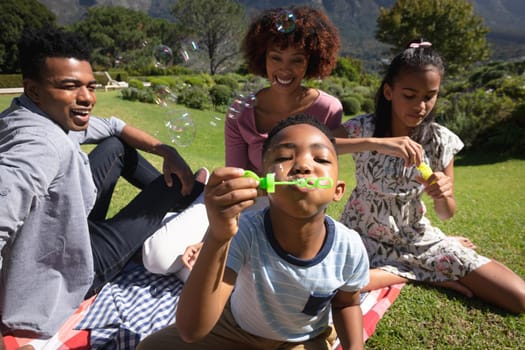 Happy african american couple with son and daughter outdoors, blowing bubbles in sunny garden. family enjoying quality free time together.