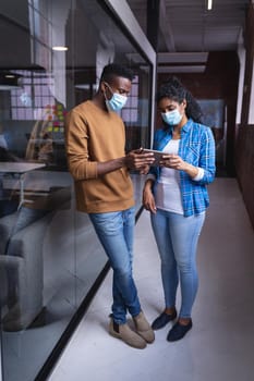 Diverse male and female colleagues at work discussing and looking at tablet wearing face masks. independent creative design business during covid 19 coronavirus pandemic.