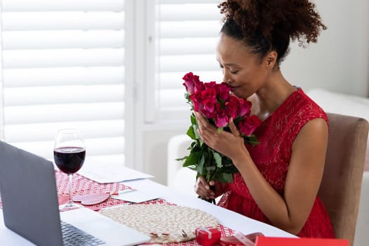 African american woman smelling flower bouquet on videocall at home. distant celebration of valentines day online and dating concept