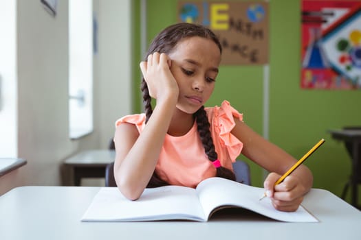 Mixed race schoolgirl in classroom sitting at desk concentrating and writing in book. childhood and education at elementary school.