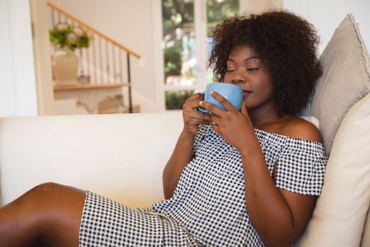 Portrait of smiling african american woman with eyes closed having tea sitting on sofa at home. domestic lifestyle, enjoying leisure time at home.