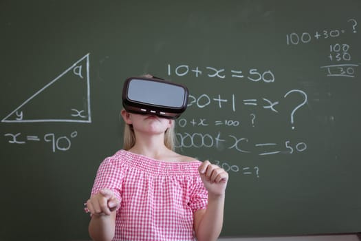 Caucasian schoolgirl standing at chalkboard in classroom wearing vr headset. childhood, technology and education at elementary school.