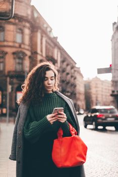 A woman on the street uses a mobile phone. online shopping. use of mobile applications. beautiful young woman with long curly dark hair in a casual coat, trendy green sweater and red handbag