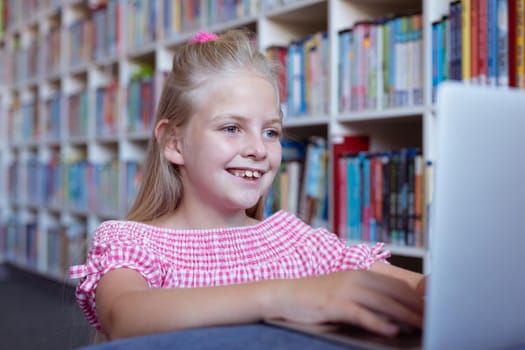 Smiling caucasian schoolgirl at desk in school library using laptop. childhood, technology and education at elementary school.