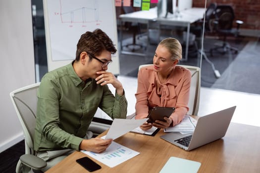 Diverse male and female colleague sitting at table with laptop and paperwork discussing. working in business at a modern office.