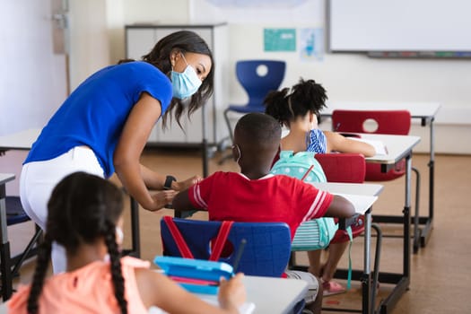 African american female teacher wearing face mask teaching african american boy in class at school. education back to school health safety during covid19 coronavirus pandemic