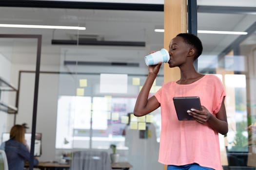 African american businesswoman standing at corridor drinking coffee using digital tablet. business person in a modern office.