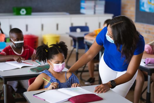 African american female teacher wearing face mask teaching african american girl in class at school. education back to school health safety during covid19 coronavirus pandemic
