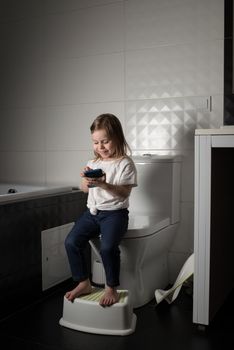 A girl dressed in a blue jeance and white t-shirt playing with a mobile phone in the bathroom.