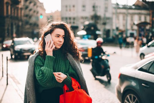 A woman on the street uses a mobile phone. online shopping. use of mobile applications. beautiful young woman with long curly dark hair in a casual coat, trendy green sweater and red handbag