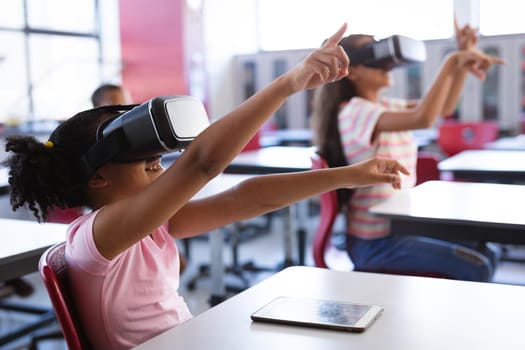 African american girl wearing vr headset gesturing while sitting on her desk in the class at school. school and education concept