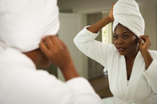 African american woman in bathroom wearing bathrobe, looking in mirror putting towel on head. health, beauty and wellbeing, spending quality time at home.