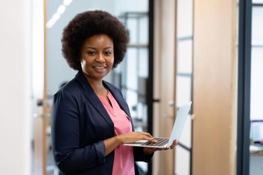 Happy african american businesswoman standing in corridor using laptop looking to camera. business in a modern office.