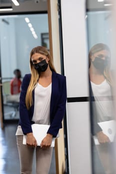 Caucasian businesswoman wearing mask standing in corridor holding laptop looking to camera. business in a modern office during covid 19 coronavirus pandemic.