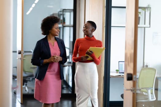 Happy african american female business colleagues walking through corridor holding documents talking. business in a modern office.