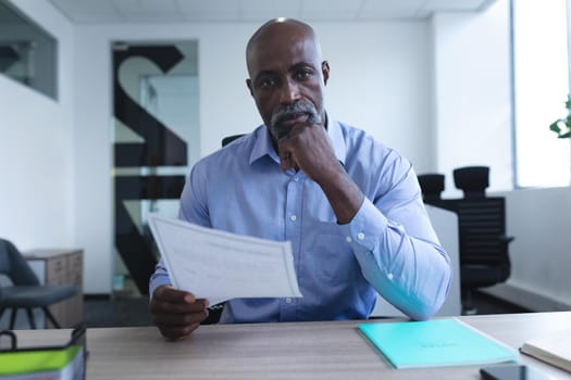 Portrait of african american businessman sitting at desk and holding documents, having video call. online meeting, working in isolation during quarantine lockdown.