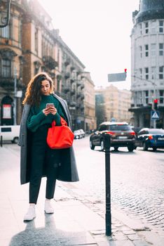 A woman on the street uses a mobile phone. online shopping. use of mobile applications. beautiful young woman with long curly dark hair in a casual coat, trendy green sweater and red handbag