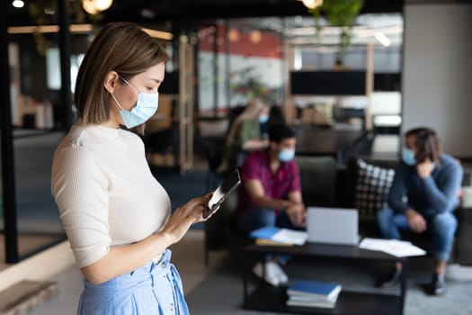 Asian businesswoman wearing face mask using tablet in creative office. social distancing health protection hygiene in workplace during covid 19 pandemic