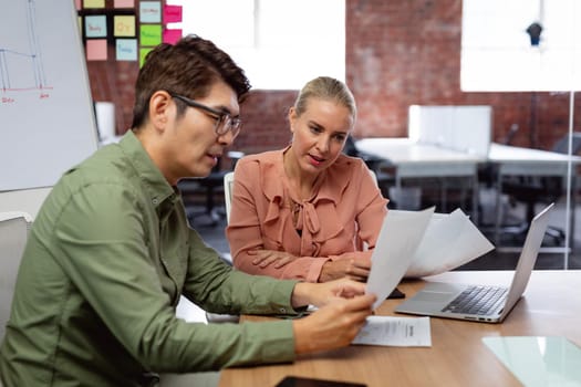 Diverse male and female colleague sitting at table with laptop and paperwork discussing. working in business at a modern office.