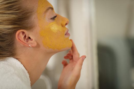 Caucasian woman in bathroom wearing bathrobe, looking in mirror and applying face mask. health, beauty and wellbeing, spending quality time at home.