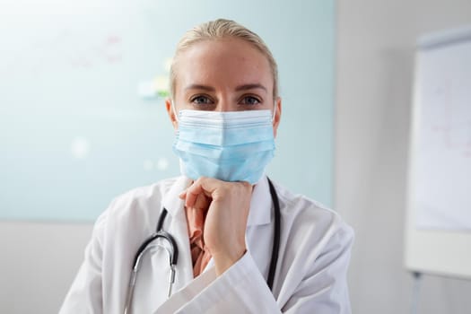 Caucasian female doctor wearing face mask sitting at desk in office listening during video call. telemedicine, online medical and healthcare services during coronavirus covid 19 pandemic.