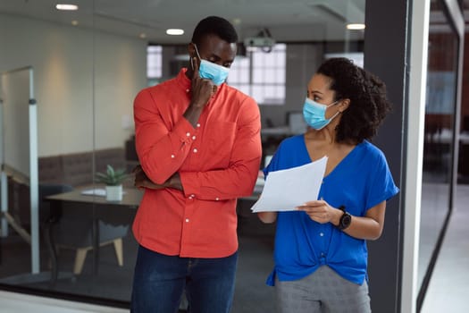 Diverse male and female colleague in face masks looking at paperwork and discussing in office. working in business at a modern office during coronavirus covid 19 pandemic. .
