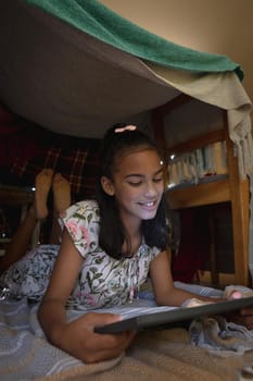 Happy african american girl at home, lying in blanket fort and using tablet. childhood with technology, spending free time at home.