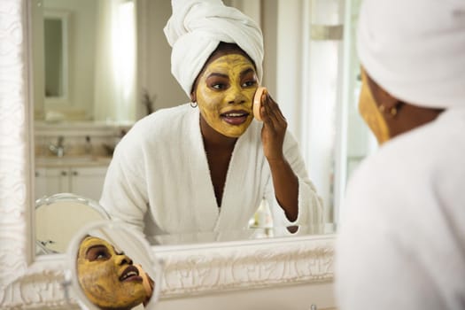 Happy african american woman in bathroom with towel on head, looking in mirror applying face mask. health, beauty and wellbeing, spending quality time at home.