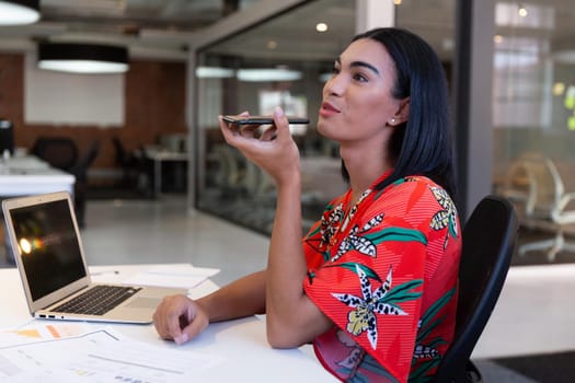 Portrait of mixed race businesswoman sitting in front of laptop having a call on smartphone. independent creative design business.