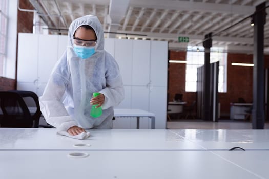 Cleaner wearing ppe suit, glasses and mask disinfecting office workspace, wiping desks. hygiene in business at a modern office during coronavirus covid 19 pandemic.