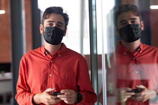 Portrait of caucasian businessman wearing face mask standing in corridor using smartphone. independent creative business during covid 19 coronavirus pandemic.